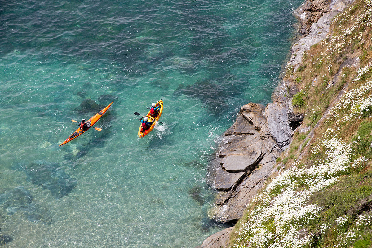 birds-eye view of two sea kayakers paddling across crystal clear turquoise waters on a trip with cornish rock tors