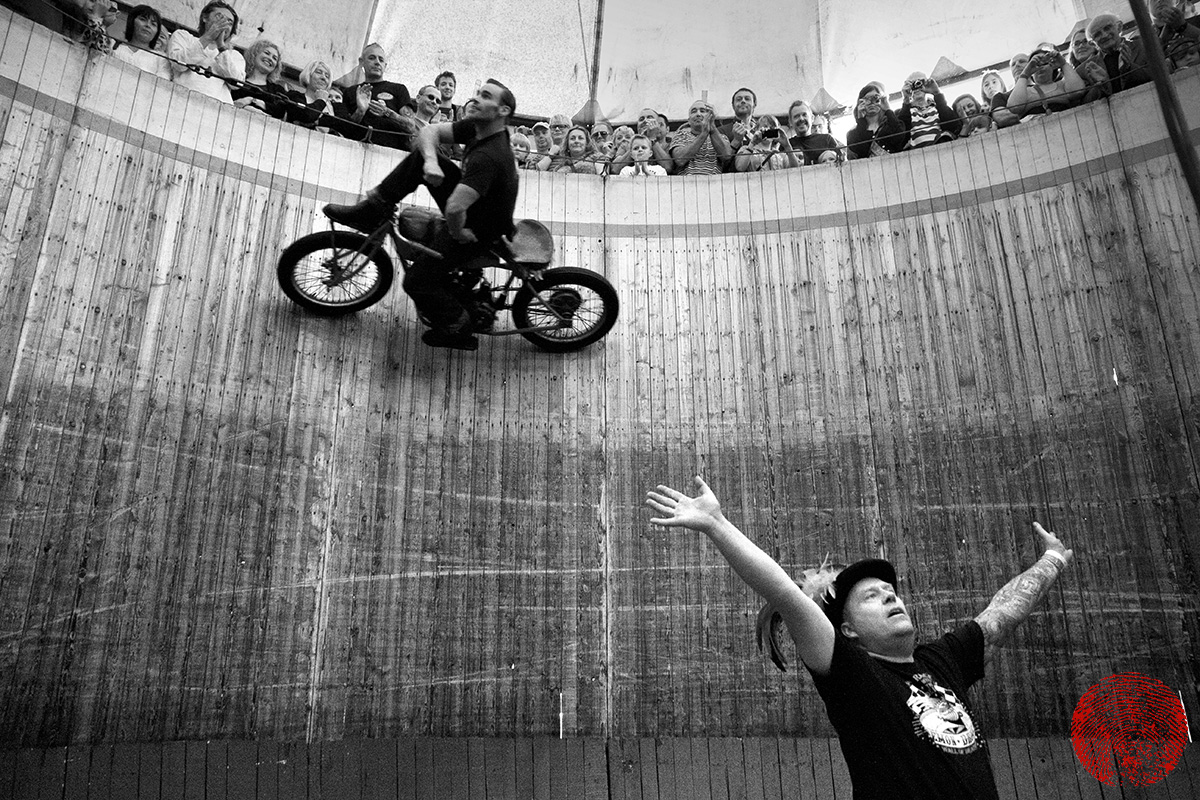 the duke riding a vintage indian motorcycle sat on the handlebars on the demon drome wall of death with the compere beneath him